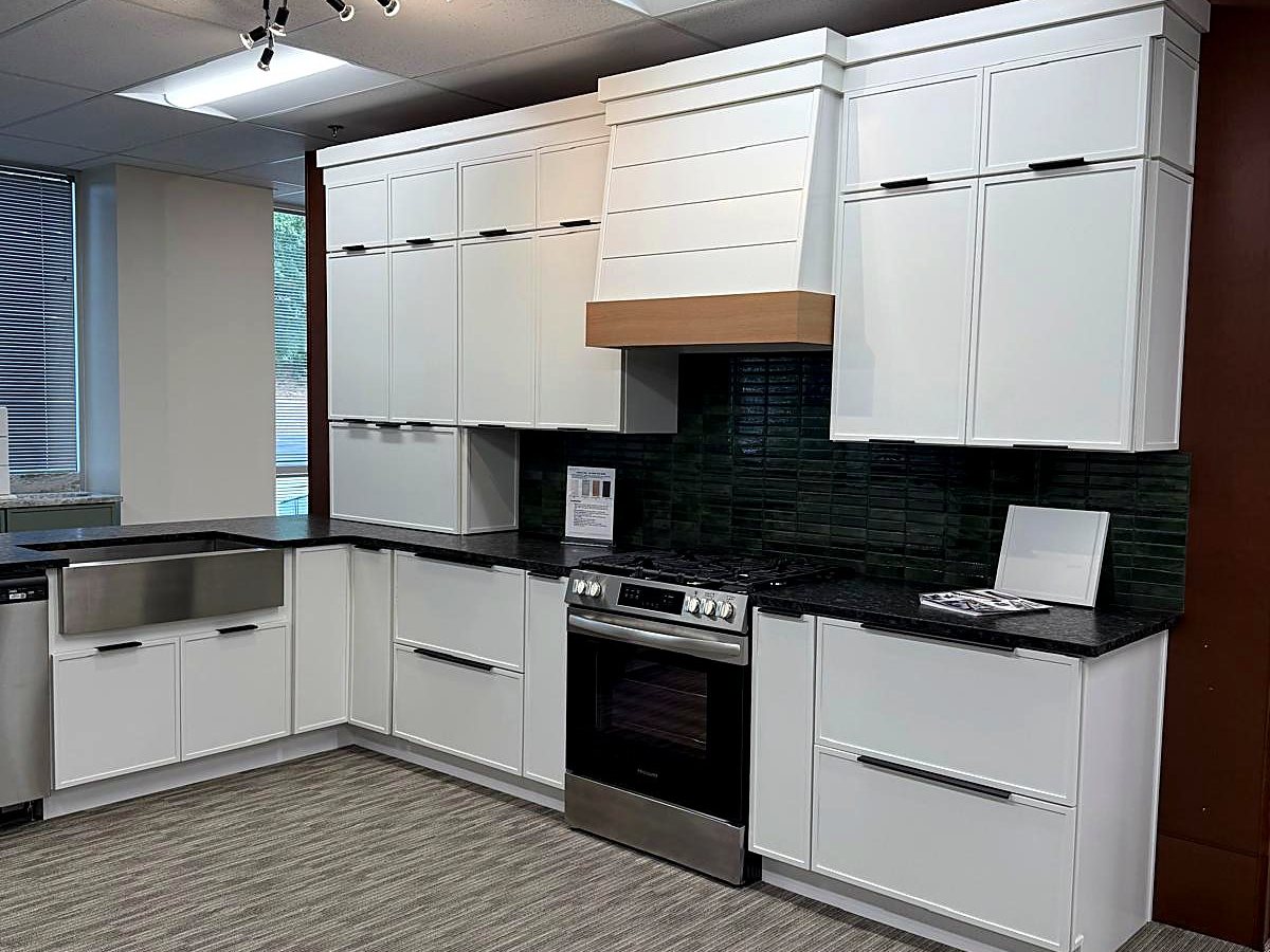 White Slim Shaker display kitchen with a row of cabinets stacked above the uppers, a shiplap range hood with natural wood trim and a dark green tile backsplash, at the MNK Cabinet showroom in Norcross